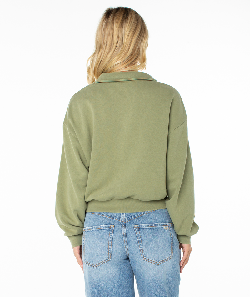 A person with long, wavy blonde hair is wearing the Roxy Half Moon Top in a light green cotton blend and blue jeans, standing with their back to the camera against a white background. The cozy fabric of the top exudes lounge comfort.