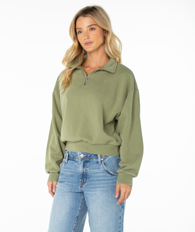 A woman wearing the Roxy Half Moon Top in light green and blue jeans stands against a plain white background, looking slightly to the side with a neutral expression. The cotton blend fabric of the Half Moon Top enhances lounge comfort while maintaining a stylish, relaxed look.