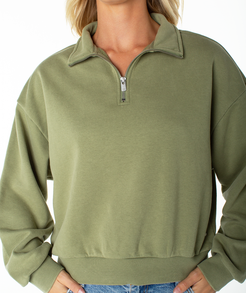 A woman wearing the Roxy Half Moon Top—a light olive green quarter zip crafted from a soft cotton blend with a collar and long sleeves—paired with blue jeans. The image is cropped from shoulders to hips, highlighting the top’s effortless lounge comfort.