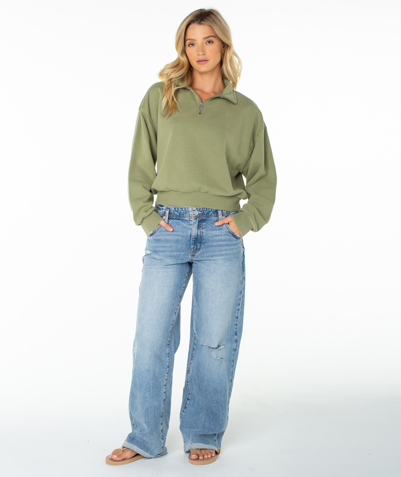 A woman wearing the Roxy Half Moon Top in green cotton blend and light blue wide-leg jeans stands with her hands in her pockets, sandals on her feet, against a plain white background.