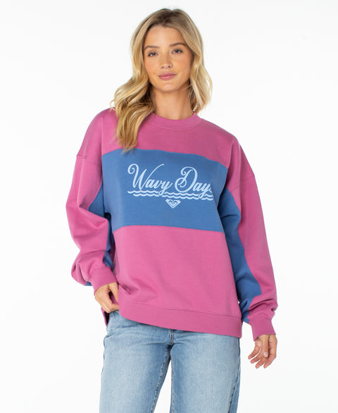 A woman with long blonde hair wears the Roxy Lineup Colorblock Crew, an oversized, ultra-soft cotton crewneck sweatshirt in a Red Violet Wavy Day color combination. She pairs it with light blue jeans and stands facing forward against a white background.