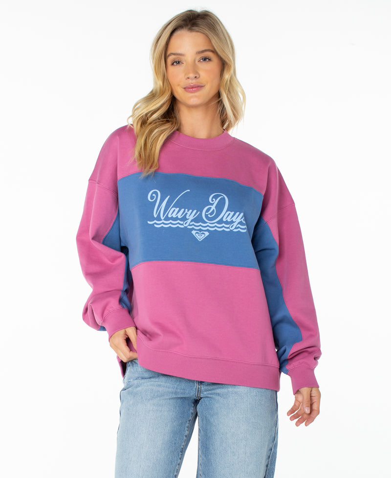 A woman with long blonde hair wears the Roxy Lineup Colorblock Crew, an oversized, ultra-soft cotton crewneck sweatshirt in a Red Violet Wavy Day color combination. She pairs it with light blue jeans and stands facing forward against a white background.