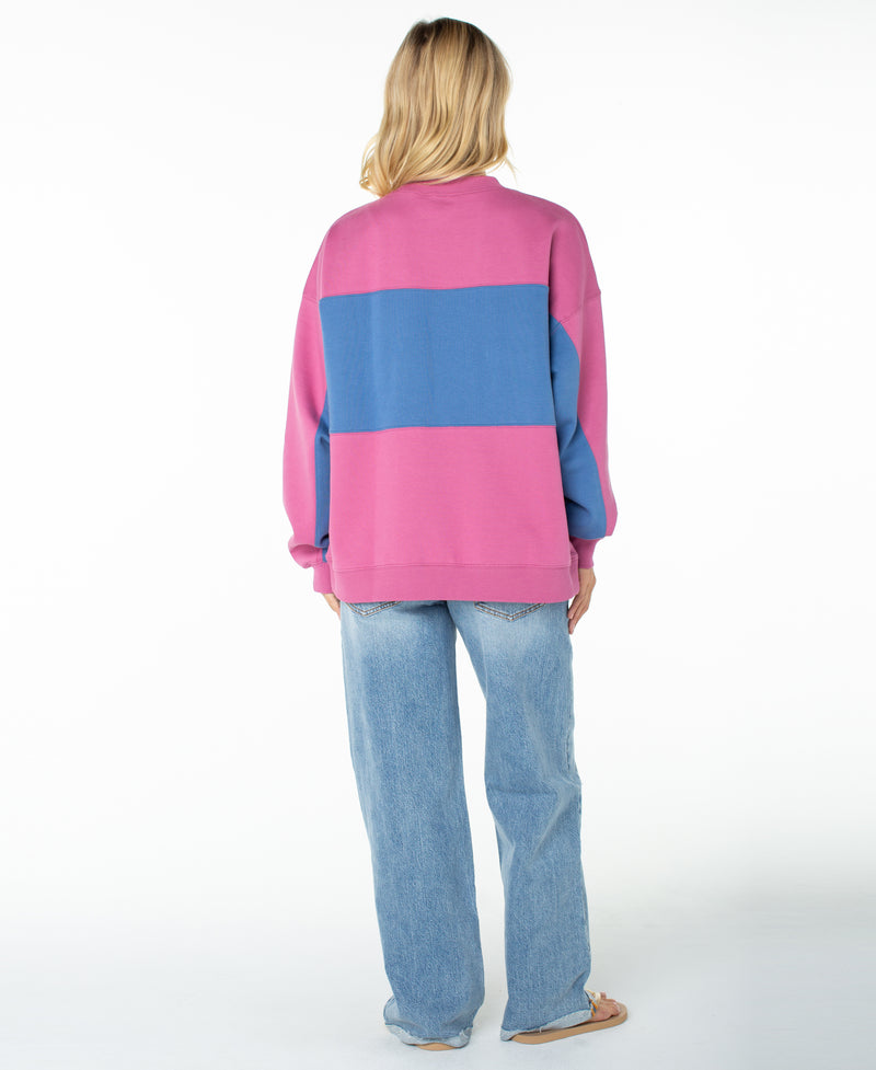 A person with long blonde hair stands facing away from the camera against a white background, wearing the Roxy Lineup Colorblock Crew—an oversized pink and blue sweater crafted from ultra-soft cotton—paired with light blue jeans and sandals.