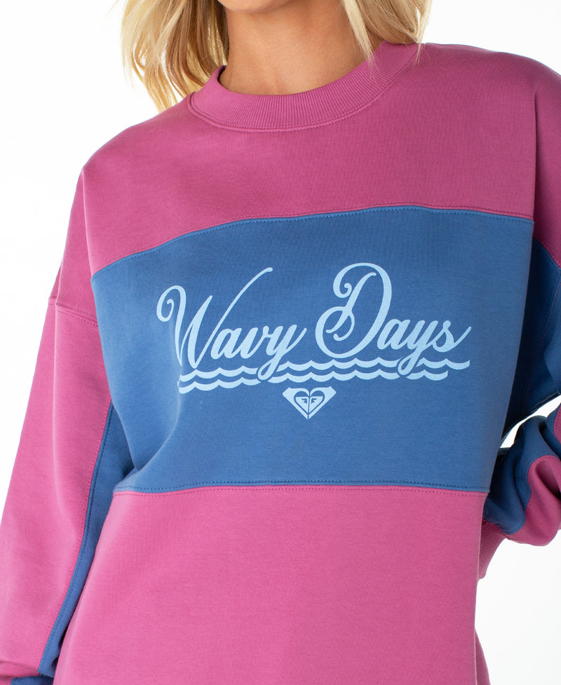 A person wears the Roxy Lineup Colorblock Crew, an oversized crewneck featuring a pink and blue color block design, “Wavy Days” text with a small logo beneath, crafted from ultra-soft cotton; shown in a shoulder-to-waist photograph.