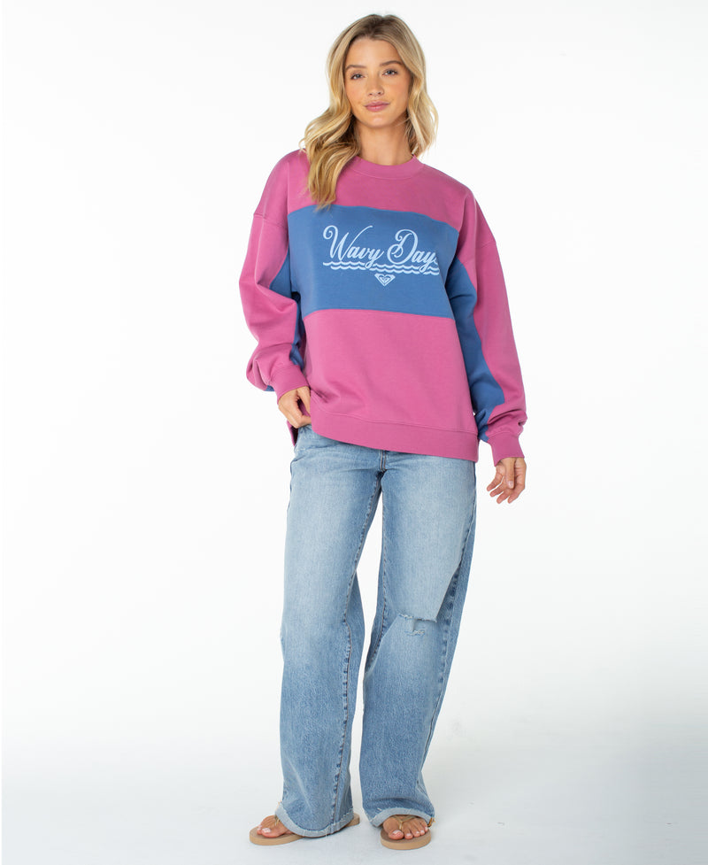 A woman with long blonde hair stands against a white background, looking at the camera with a relaxed expression. She is wearing the Roxy Lineup Colorblock Crew—an ultra-soft cotton sweatshirt—and light blue jeans.