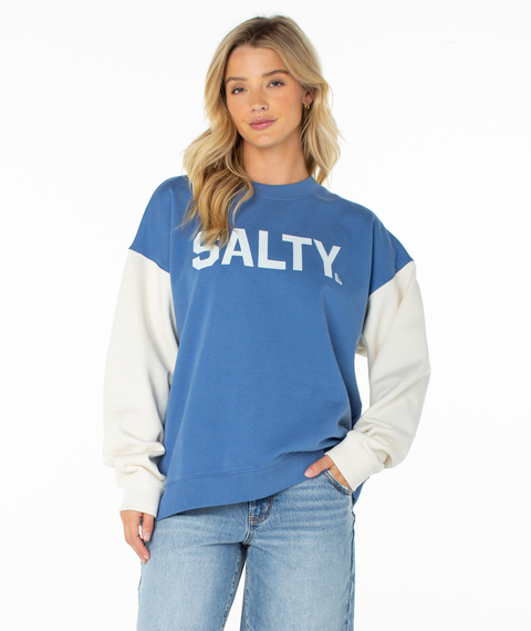 A woman with long blonde hair wears the Roxy Lineup Contrast Crew in Dutch Blue Salty College, featuring color block white sleeves and "SALTY." on the front, paired with light blue jeans against a plain white background.