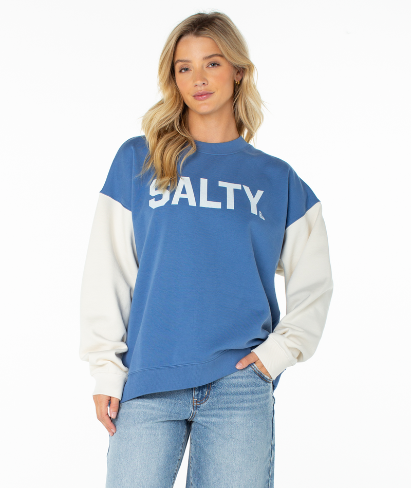 A woman with long blonde hair wears the Roxy Lineup Contrast Crew in Dutch Blue Salty College, featuring color block white sleeves and "SALTY." on the front, paired with light blue jeans against a plain white background.