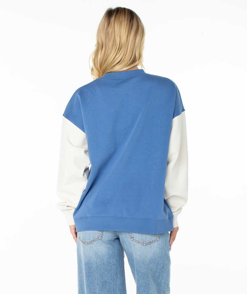 A person with long blonde hair is shown from the back, wearing the Roxy Lineup Contrast Crew—an oversized, ultra-soft color block sweatshirt—and light blue jeans, set against a white background.