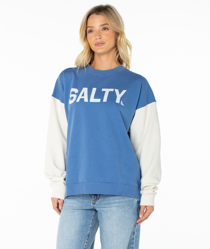 A woman wearing the Roxy Lineup Contrast Crew, an ultra-soft blue sweatshirt with "SALTY" in white letters, color block white sleeves, and blue jeans stands against a plain white background.
