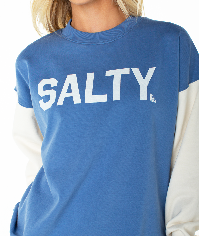 A woman wears the Roxy Lineup Contrast Crew, a color block blue oversized crewneck with white sleeves and "SALTY" in bold white letters across the chest. Only her lower face is visible. Made from ultra-soft cotton for all-day comfort.