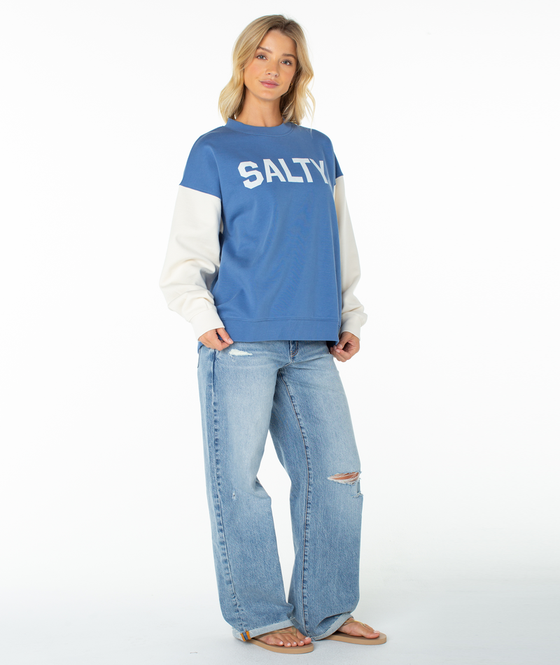 A woman with blonde hair and a relaxed expression wears the Roxy Lineup Contrast Crew, an oversized crewneck blue sweatshirt with SALTY text, light color-blocked long sleeves, ultra-soft cotton fabric, paired with ripped blue jeans and sandals.