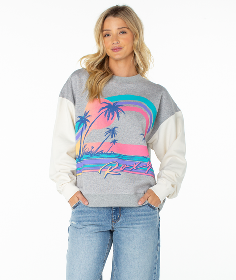 A woman in a Roxy Lineup Contrast Crew sweatshirt in Heather Gray Heatwave, featuring a colorful sunset and palm trees, paired with blue jeans, stands against a white background.