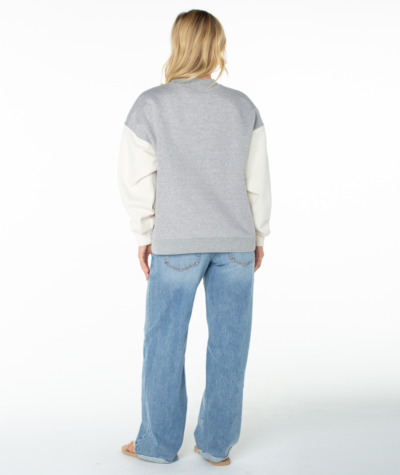 A person with blonde hair stands facing away, wearing the Roxy Lineup Contrast Crew—an oversized gray and white color block sweatshirt—paired with light blue wide-leg jeans and sandals against a plain white background.