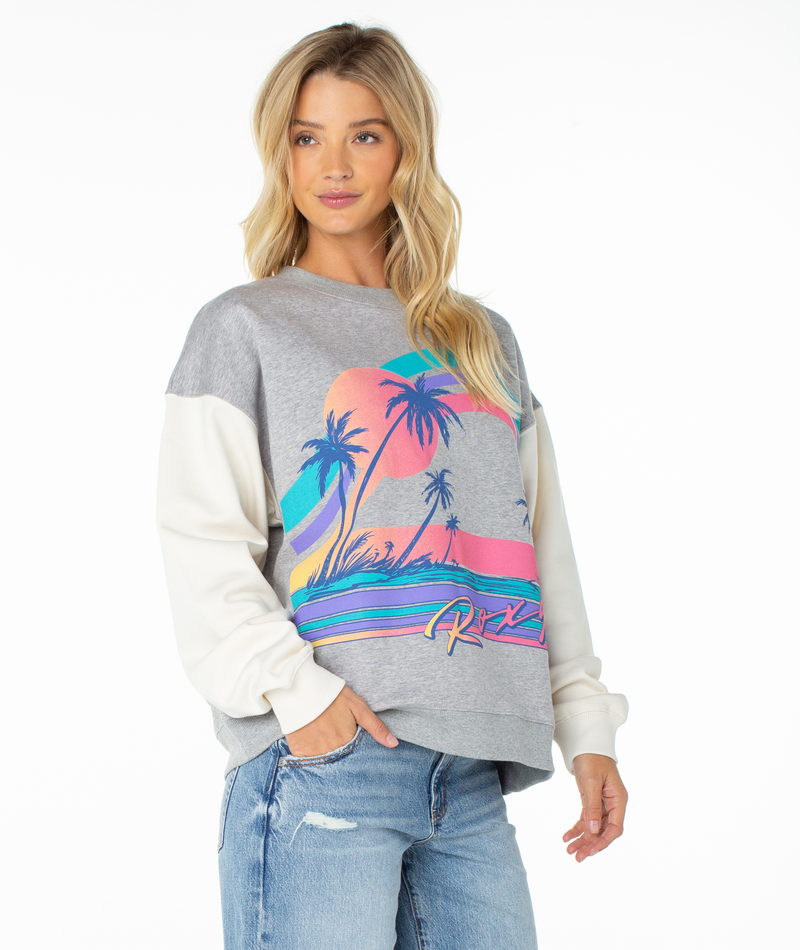A woman with long blonde hair wears the Roxy Lineup Contrast Crew, an oversized gray and white sweatshirt with a colorful tropical sunset and palm trees, paired with light blue ripped jeans, standing against a white background.