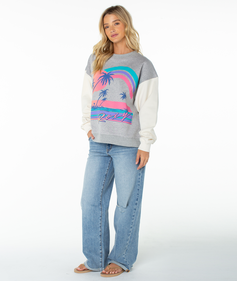 A woman with long blonde hair stands against a white background wearing the Roxy Lineup Contrast Crew, an oversized grey sweatshirt with colorful palm tree graphics, paired with light blue wide-leg jeans and sandals. She has a relaxed expression.