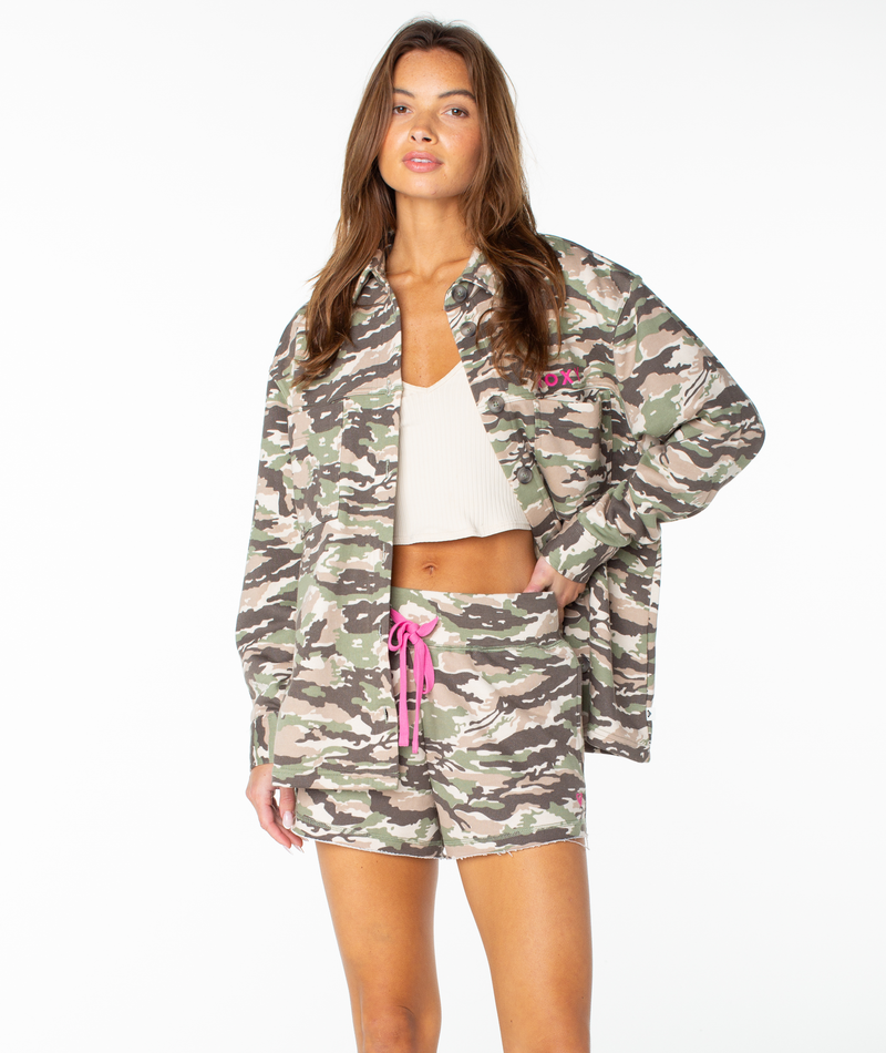 A woman models the Roxy Dawn Light Button Up jacket and matching shorts, crafted from a soft cotton blend with a light camouflage print and finished with a bright pink drawstring waist. She wears them over a white crop top, standing against a plain white background and looking at the camera with one hand in her pocket.