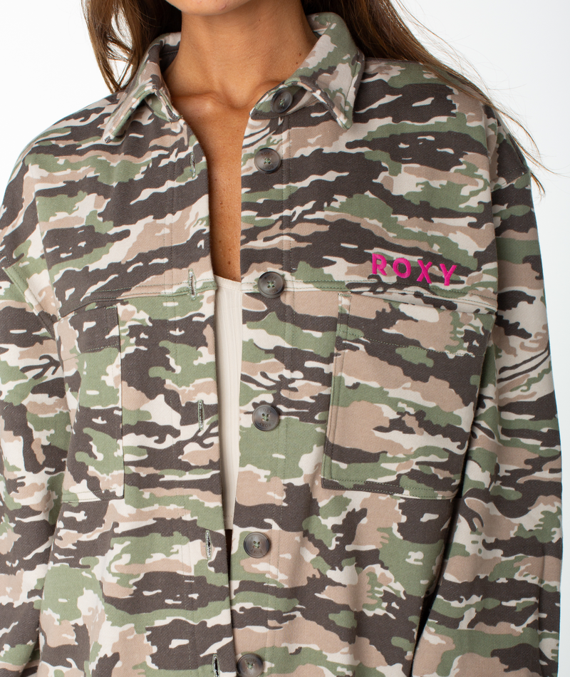 A woman wears the Roxy Dawn Light Button Up—a camo print shirt with two chest pockets, made from a soft cotton blend. “ROXY” is embroidered in pink on the left pocket. Only the lower half of her face and torso are visible.