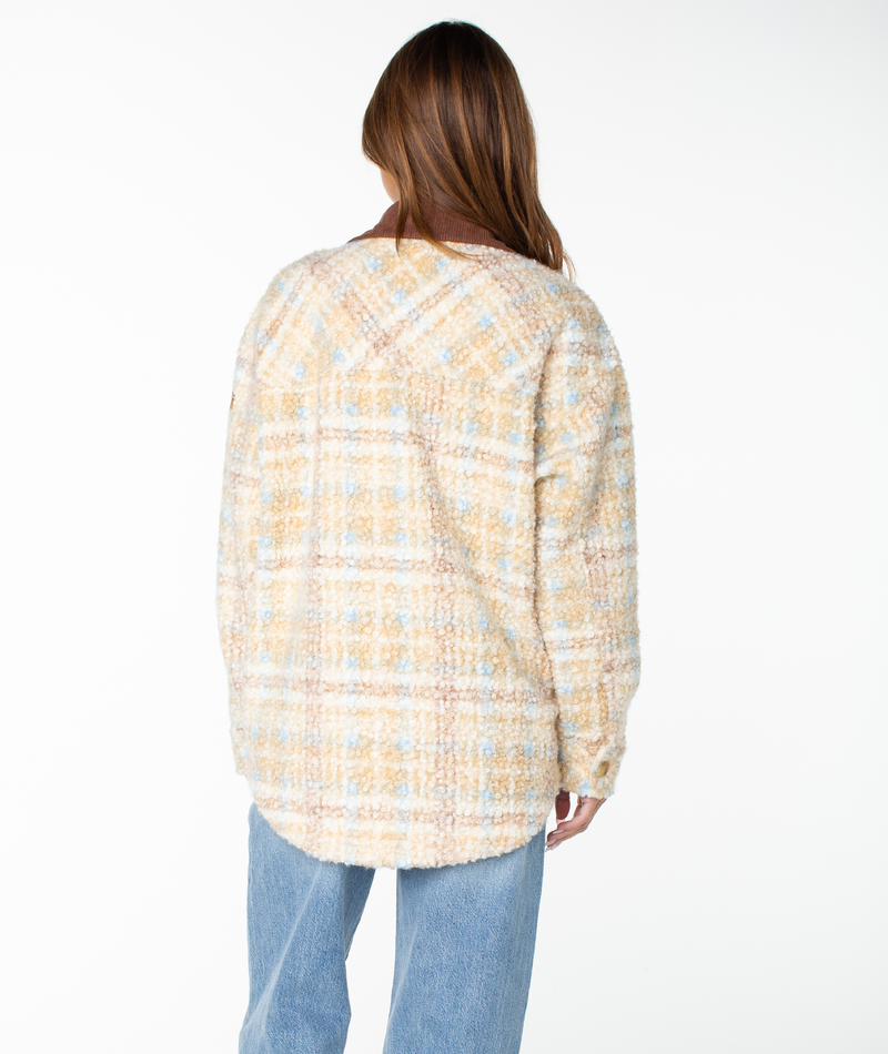 A person with long brown hair wearing the Roxy Overlander Jacket—a flannel-inspired pastel plaid piece that’s a perfect fall staple—and light blue jeans stands facing away from the camera against a white background.