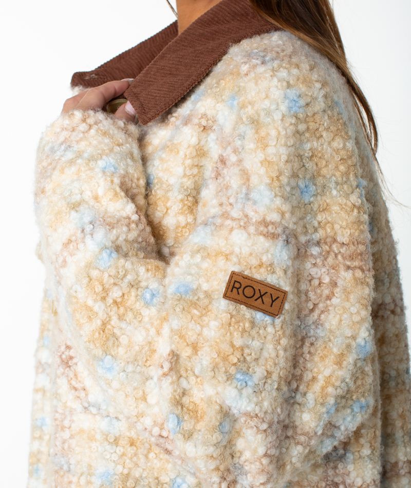A person wears the Roxy Overlander Jacket—a fuzzy, patterned beige and blue piece featuring a brown collar. The flannel-inspired design enhances its cozy appeal, making it an ideal fall staple. A ROXY patch appears on the sleeve, and the person’s face is not visible.