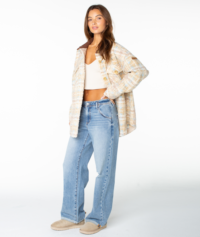 A woman stands in a white studio, wearing the Overlander Jacket by Roxy layered over a white crop top, paired with light blue wide-leg jeans and beige slip-on shoes. Her long hair and relaxed, confident expression capture the essence of an effortless fall staple look.