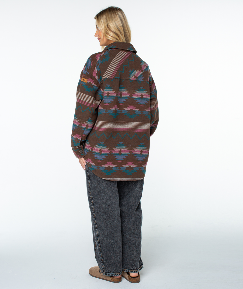 A person wearing a Roxy Overlander Jacket in an oversized brown button-up style with a teal, pink, and white geometric pattern, paired with black jeans and brown slip-on shoes.