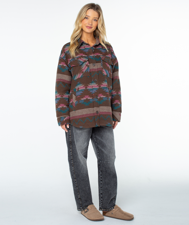 A woman wears an oversized Roxy Overlander Jacket in Chestnut Baja Blanket with a southwestern geometric pattern, grey jeans, and tan slip-on shoes.