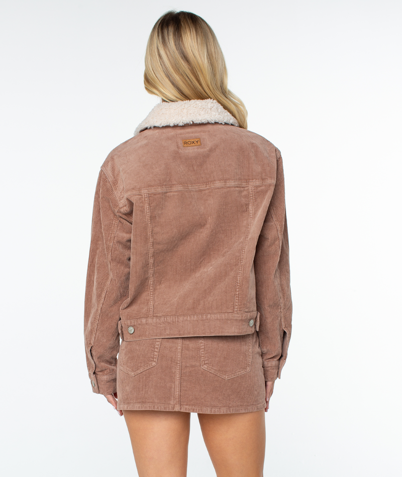 A person with long blonde hair models the Roxy Big Smoke Jacket, featuring a corduroy blend and white sherpa collar, paired with a matching skirt, standing back to the camera against a plain white background.