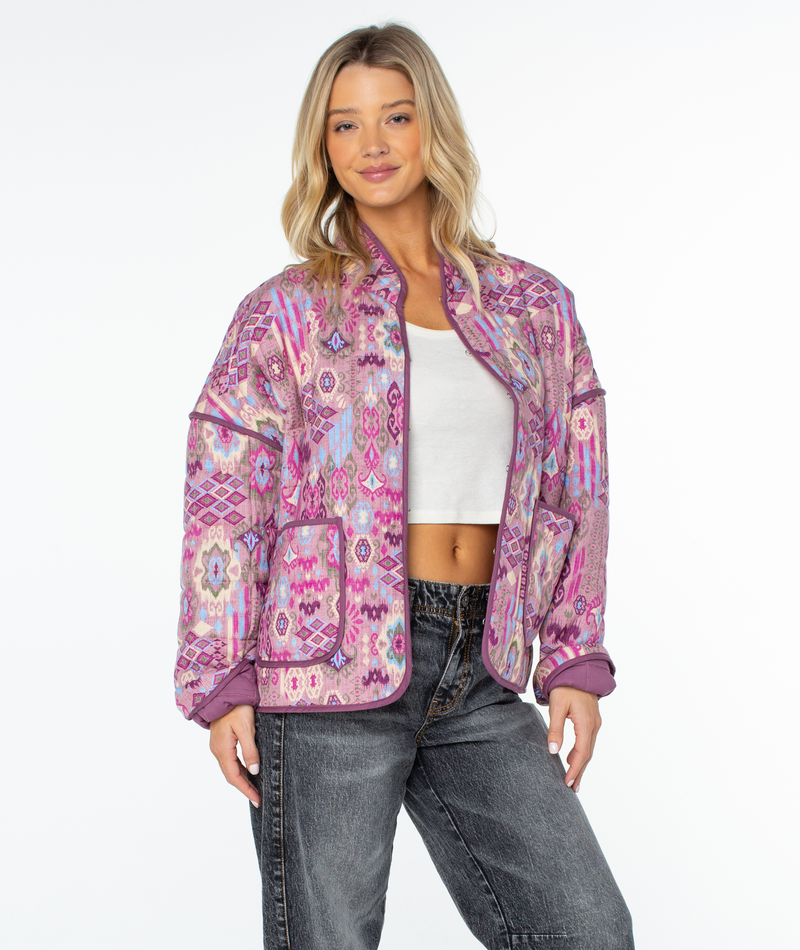 A woman wears a relaxed fit Roxy Solana Jacket in pink, featuring a quilted geometric design and purple trim over a white crop top and dark jeans.