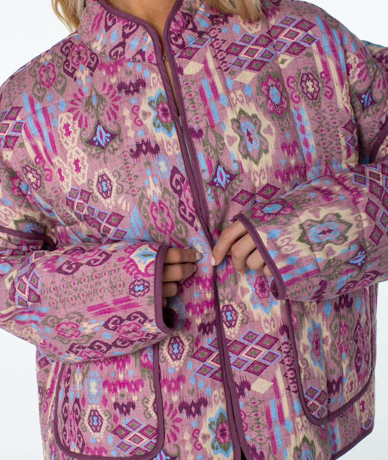 A person wearing a relaxed-fit Roxy Solana Jacket with a quilted design featuring a pink, blue, and purple geometric print and dark purple trim.