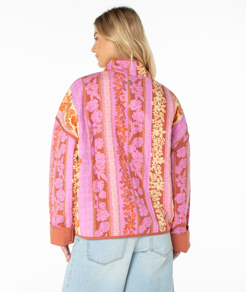 A woman with blonde hair is seen from the back wearing the Roxy Solana Quilted Jacket, featuring a cozy cotton blend with pink, orange, and yellow floral and striped patterns, paired with light blue jeans against a white background.