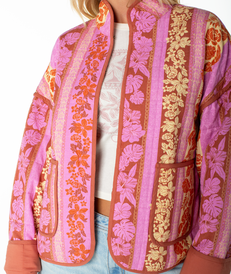 A person wears the Roxy Solana Quilted Jacket, featuring vibrant pink, orange, and yellow floral and leaf patterns, large pockets, and a high collar over a white shirt and light blue jeans.