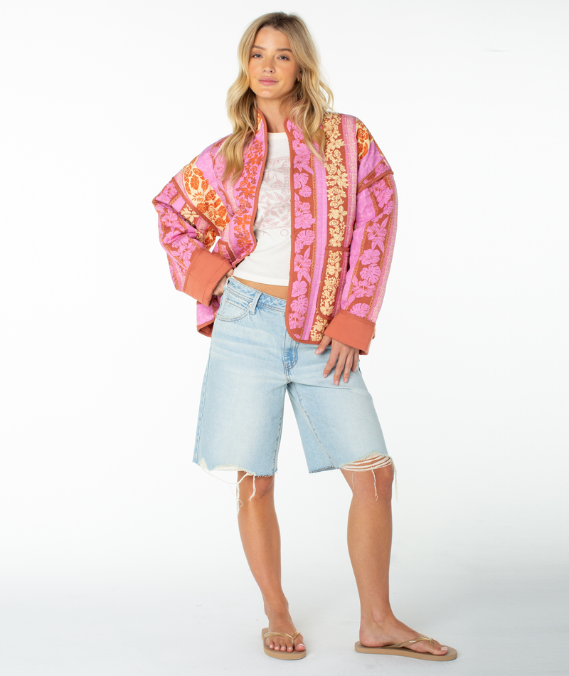 A woman with long blonde hair poses against a white background in the Roxy Solana Quilted Jacket in pink and orange, paired with a white top, light blue knee-length ripped denim shorts, and tan flip-flops.