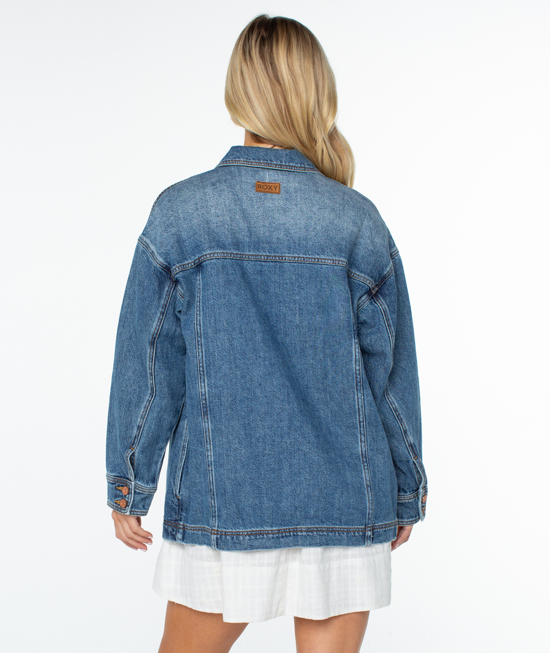 Person viewed from behind wearing an oversized blue denim Roxy Lost Treasure Jacket with a brown brand patch at the back of the collar.