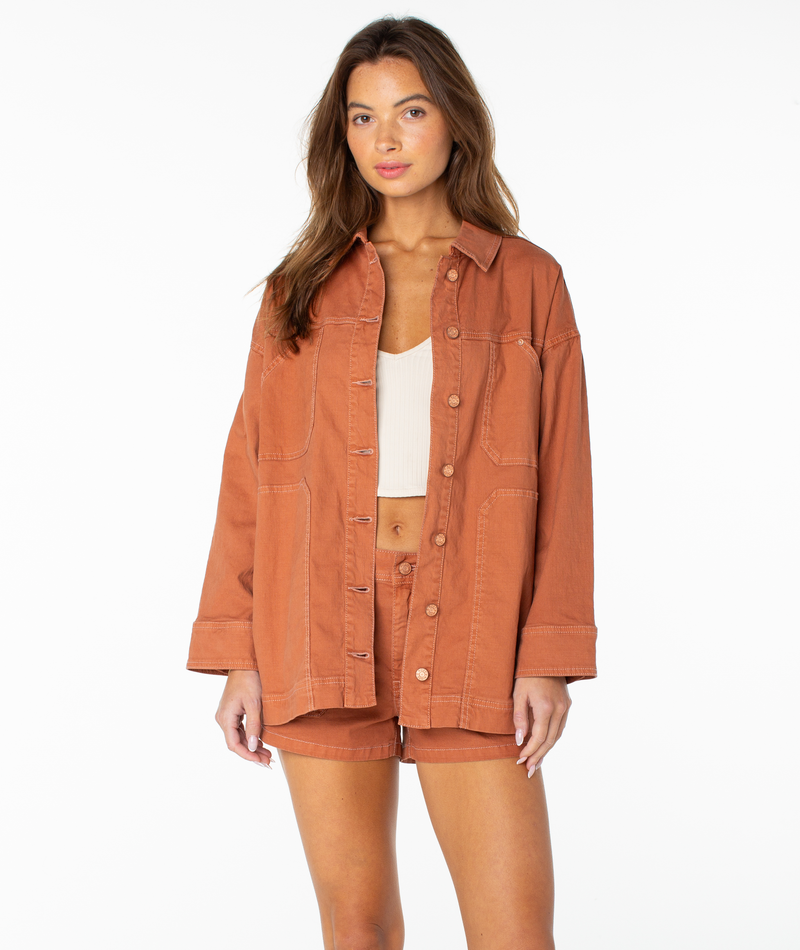 A woman with long brown hair and a neutral expression stands against a white background, wearing the Roxy Lost Treasure Jacket in Chutney Solid paired with matching shorts over a white ribbed crop top.