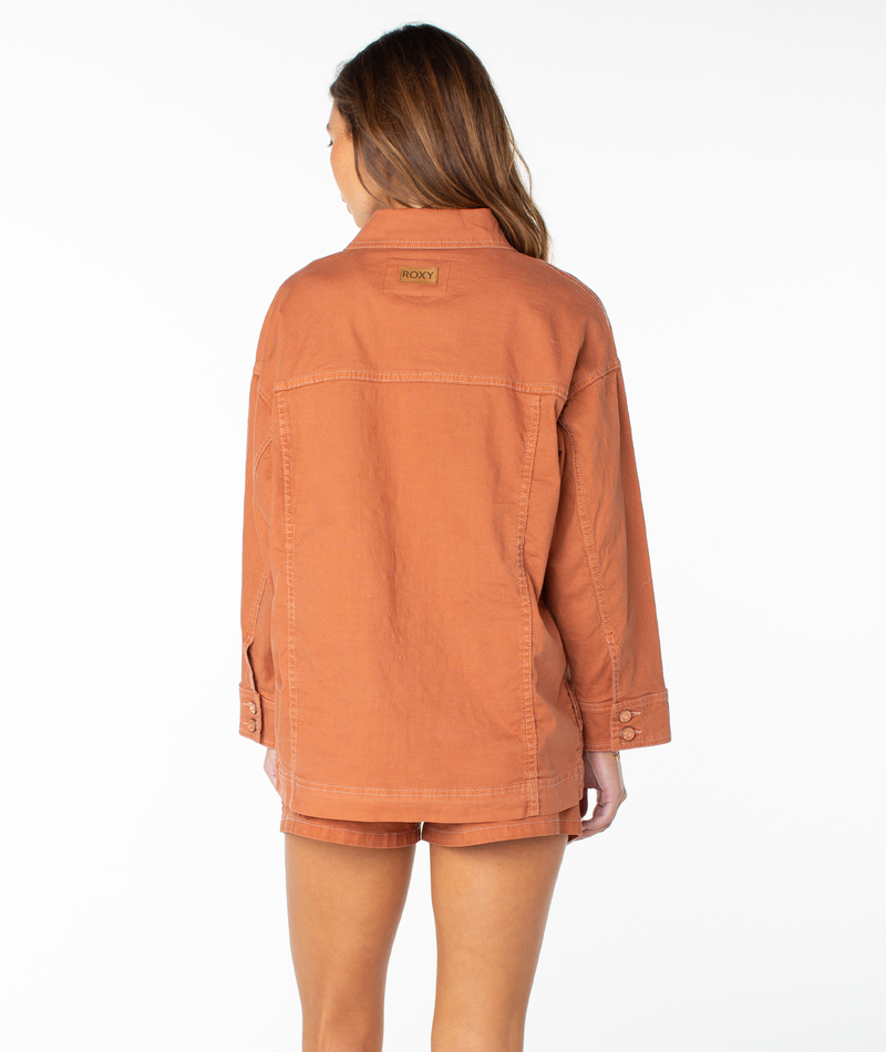 A woman stands facing away, wearing an oversized orange Roxy Lost Treasure Jacket paired with matching shorts. The background is white and minimal.