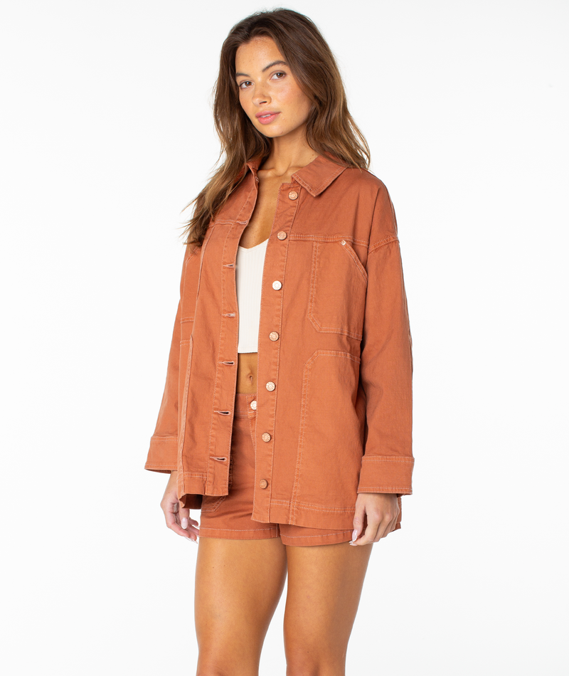 A woman wearing the Roxy Lost Treasure Jacket in a rust hue with matching shorts over a white crop top stands against a plain white background.