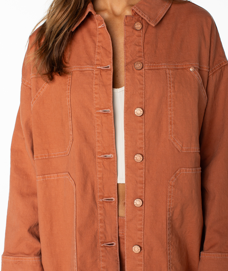 A person wearing the Roxy Lost Treasure Jacket, a rust-colored, vintage-inspired piece with oversized fit, large front pockets, and visible stitching, layered over a white top. The person’s face is not visible.
