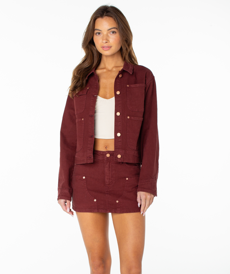A woman stands facing forward against a plain white background, wearing a Roxy Reel Life Jacket in Port Solid with oversized pockets and a matching maroon skirt. She has long brown hair and layers a white crop top underneath the jacket.