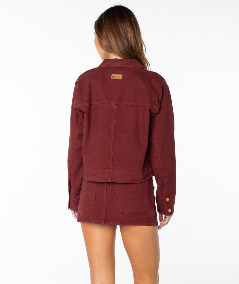 A woman stands facing away, wearing the Roxy Reel Life Jacket—a burgundy cropped piece with a small logo patch on the upper back—paired with the matching mini skirt featuring oversized pockets. The background is plain white.
