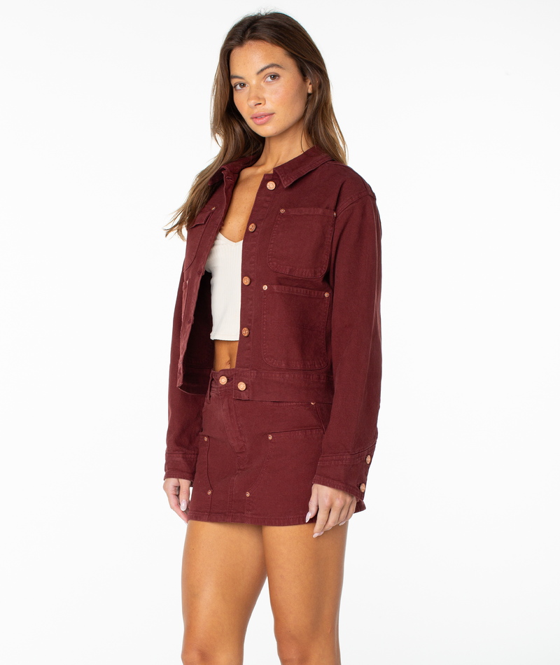 A woman with long brown hair and a neutral expression stands against a white background wearing the maroon Roxy Reel Life Jacket, which features oversized pockets, paired with a matching skirt over a white crop top.