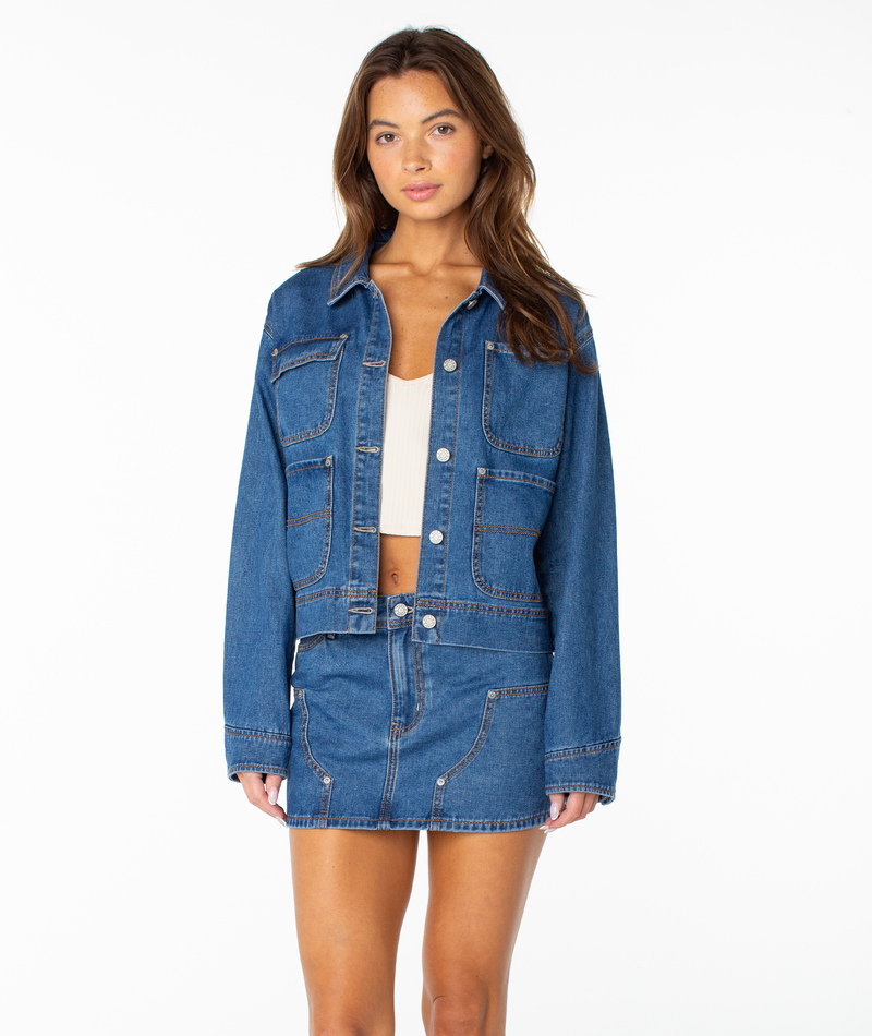 A woman stands facing forward, wearing the Roxy Reel Life Jacket in High Surf Denim Wash with oversized pockets over a white crop top and a matching denim mini skirt, her arms relaxed by her sides against a plain white background.