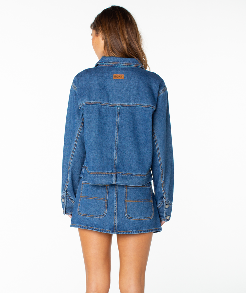 A woman stands facing away, wearing the Roxy Reel Life Jacket in a blue denim cropped fit paired with a matching denim skirt featuring back pockets and visible stitching. The background is plain white.
