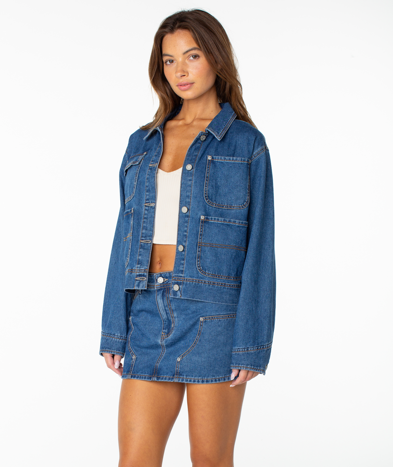 A woman wearing the Roxy Reel Life Jacket—an oversized blue denim jacket with large pockets—paired with a matching blue denim mini skirt and a white crop top, stands against a plain white background.