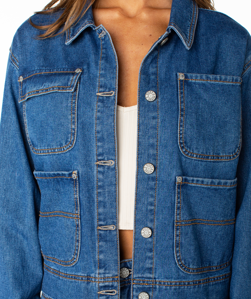 A person is seen from the torso up wearing the Reel Life Jacket by Roxy, a blue denim cropped jacket with oversized front pockets and silver buttons, styled over a white top.