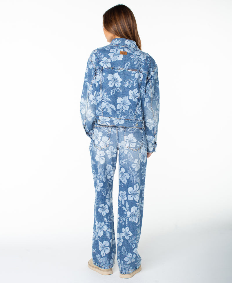 A person with long hair, seen from the back, wears the Roxy Big Tide Jacket—a blue denim piece with floral accents—paired with jeans featuring a white hibiscus pattern. They stand against a plain white background.