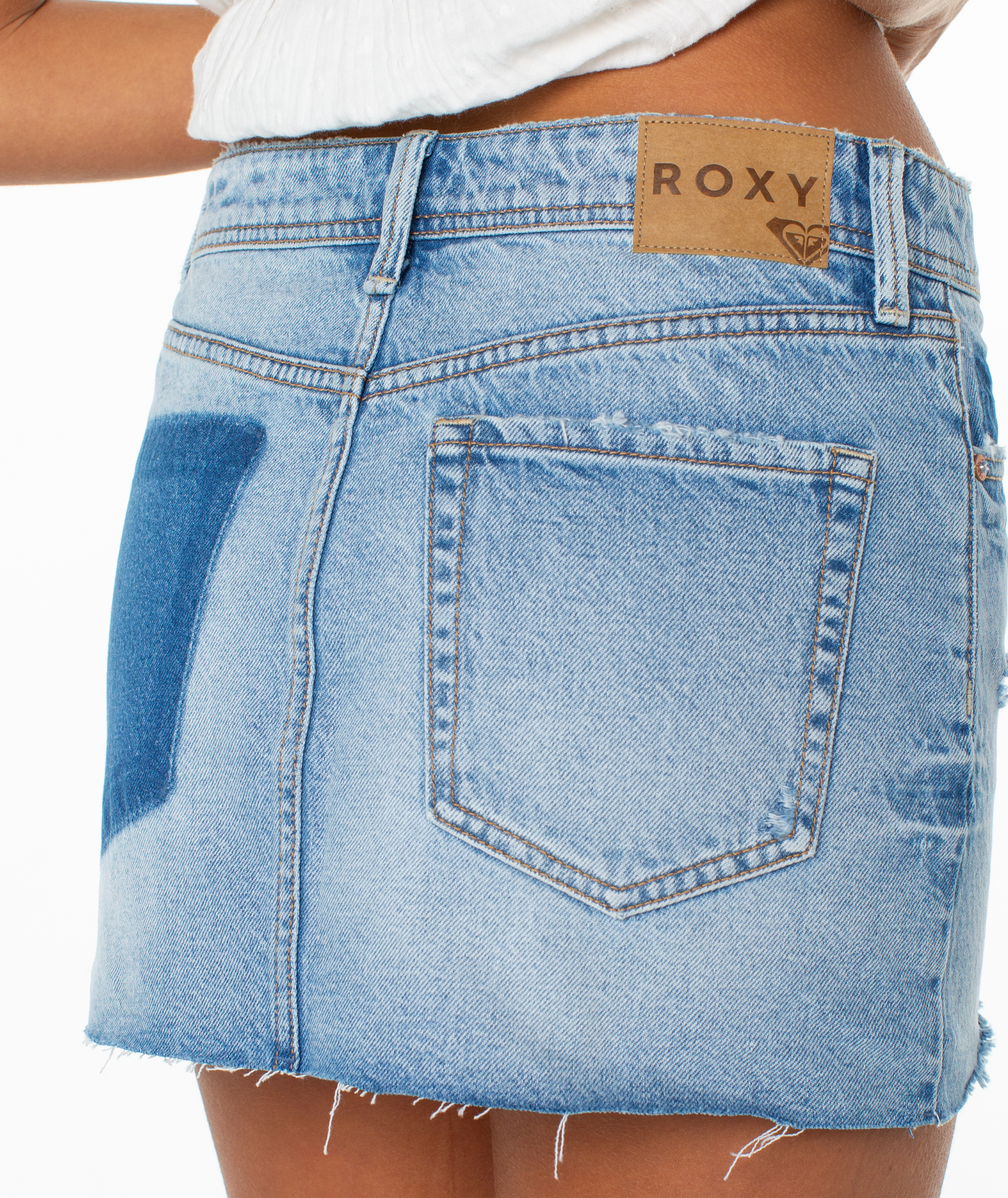 Close-up of a person wearing the Roxy Golden Horizon Skirt, a light blue denim mini with a raw hem, vintage wash, and brown Roxy leather patch above the back pocket, paired with a white top.