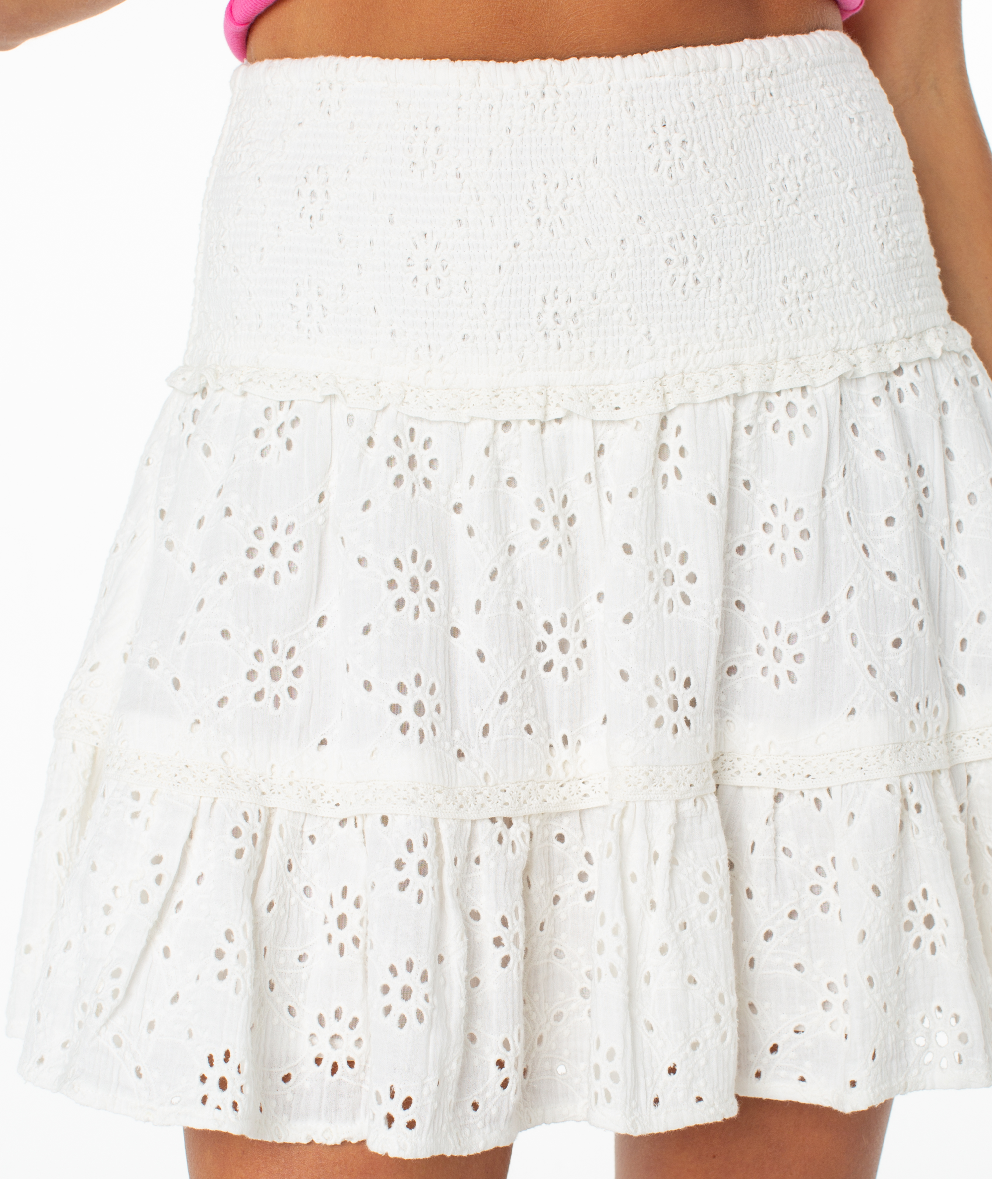 A close-up of a person wearing the Roxy Savannah Breeze Skirt, a white mini skirt with tiered ruffles and a smocked waistband. The eyelet fabric features a floral cutout pattern, styled with a pink top for summer.