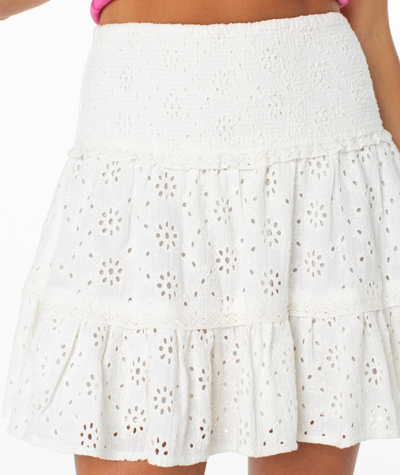 A close-up of a person wearing the Roxy Savannah Breeze Skirt, a white mini skirt with tiered ruffles and a smocked waistband. The eyelet fabric features a floral cutout pattern, styled with a pink top for summer.