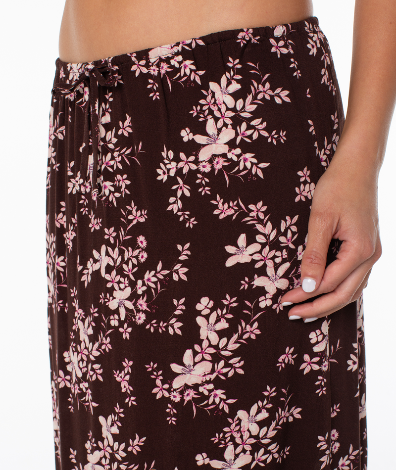 A person models the Roxy Iyla Skirt, a dark brown maxi with pink florals, against a white background. Their hand rests on the skirt, showing part of their bare midriff.
