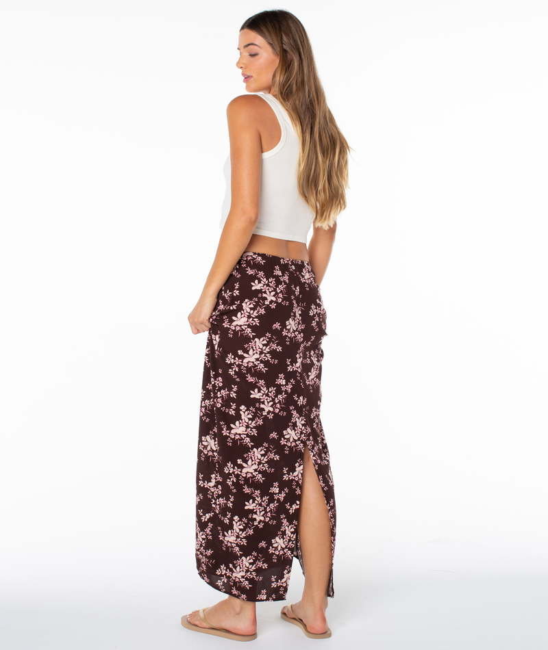 Woman wearing a white sleeveless crop top, Roxy Iyla Skirt in maroon floral with back slit, and sandals, standing sideways against a white background.